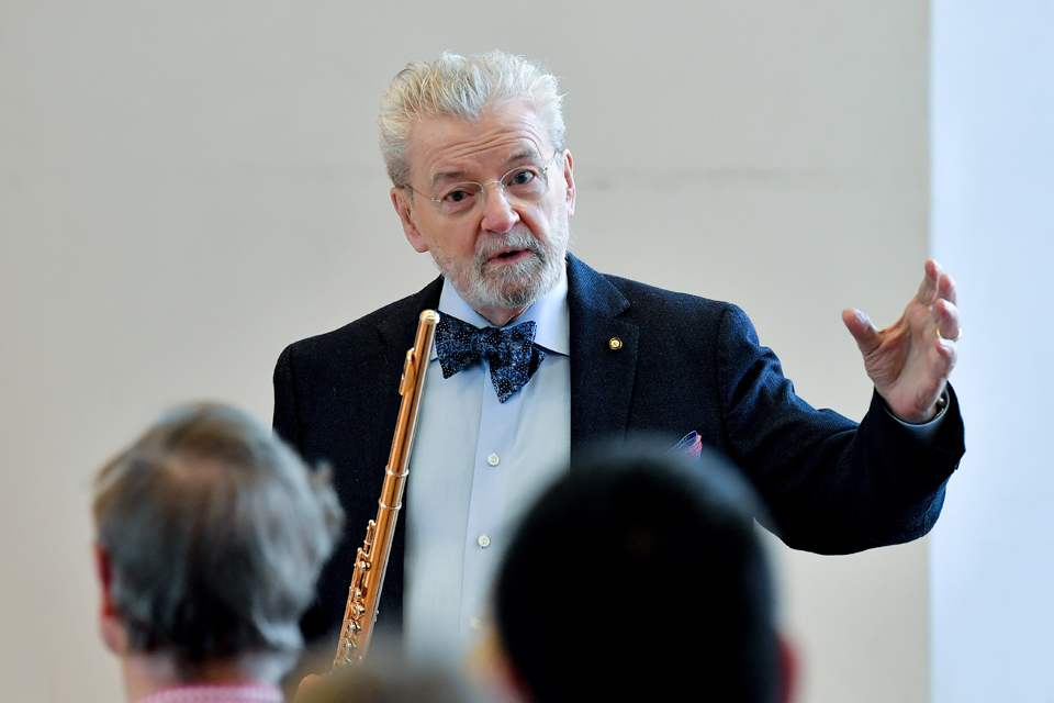 A man with white hair and glasses, Sir James Galway, teaching a class, with a golden flute in his right hand, wearing a smart suit.